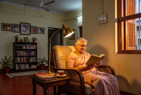 An elderly Bengali woman reading in a warm, softly lit living room, dignified and at ease